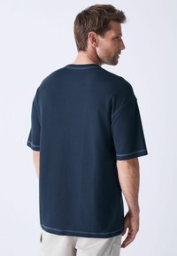 Marineblå kortærmet struktureret t-shirt med afslappet pasform, kontrasterende syninger og en rund halsudskæring, set fra ryggen.