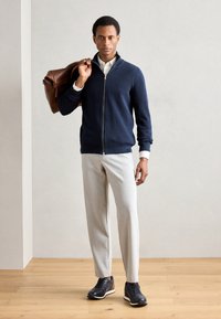 Mornarska zip-up jopica, svetlo sivi krojeni hlače in temni športni čevlji. Maneken drži rjavo usnjeno torbo, na jopici so vidne rebraste teksture.