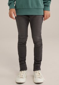 Pantalon slim gris foncé avec une texture lisse, associé à des baskets blanches à lacets et à œillets argentés.