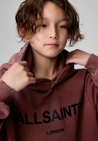 Smeđa dukserica s visokim ovratnikom, rebrastim manžetama i velikim crnim logom koji glasi "ALLSAINTS LONDON" na prednjoj strani. Mekan i teksturiran materijal.