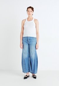 Débardeur blanc avec des côtes associé à un jean en denim bleu à jambes larges, présentant un délavage et des détails de couture subtils. Des ballerines noires complètent le look.