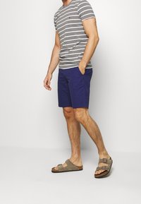 Šedobílé pruhované tričko s krátkým rukávem, námořnické šortky a hnědé sandály se dvěma popruhy. Neformální outfit s volným střihem a měkkými materiály.