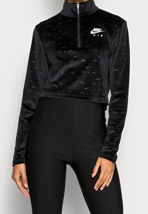 Frau trägt schwarzen Nike Air Langarm-Crop-Top mit Viertelreißverschluss und schwarze hoch taillierte Leggings, steht vor einfarbigem Hintergrund.