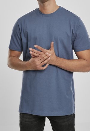 Mann trägt ein blaues kurzärmeliges T-Shirt und hält seine linke Hand mit der rechten Hand vor einem einfarbigen hellen Hintergrund fest.