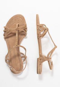 Sandales en suede tan avec plusieurs brides entrecroisées, ornées de petites clous dorés. Semelle plate, sangle de cheville ajustable, semelle intérieure texturée.