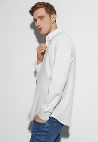 Biała koszula z długim rękawem zapinana na guziki wykonana z bawełny; posiada klasyczny kołnierzyk i detale przy mankietach, w połączeniu z niebieskimi dżinsami.