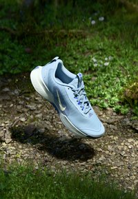 Šviesiai mėlyni Nike bėgimo batai plaukia virš akmenuoto, žolinio miško grindų, kur išsibarsčiusios žalių augalų ir mažų gėlių.