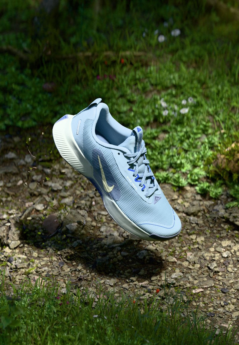 Šviesiai mėlyni Nike bėgimo batai plaukia virš akmenuoto, žolinio miško grindų, kur išsibarsčiusios žalių augalų ir mažų gėlių.