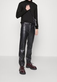 Schwarze Lederhose mit geradem Schnitt, mit Reißverschluss und Seitentaschen, kombiniert mit burgunderfarbenen Schnürschuhen und einem schwarzen Oberteil.