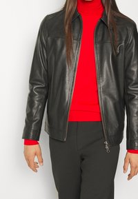 Veste en cuir noir avec fermeture éclair à l'avant, dotée d'une texture lisse et d'une coupe ajustée. Portée par-dessus un col roulé rouge vif.