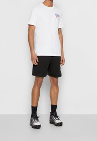 Homme portant un t-shirt graphique blanc Nike, un short noir, des chaussettes noires et des chaussures de sport noir et gris, se tenant devant un fond uni.