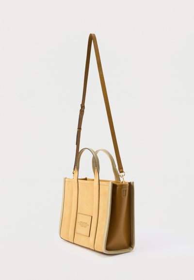 Τσάντα ώμου σε μπεζ και καφέ χρώμα με διπλές λαβές και μακρύ ρυθμιζόμενο ιμάντα ώμου, με ένα μικρό μπροστινό ταμπελάκι με κείμενο.