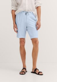 Light blue linen shorts with an elastic waistband and drawstring. Worn with a loose white shirt and dark brown sandals.