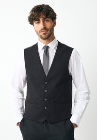 Veste noire ajustée, dotée de cinq boutons, deux poches avant et d'une texture lisse, portée sur une chemise blanche et une cravate grise.