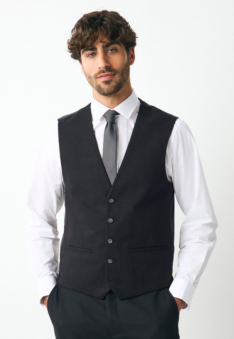 Veste noire ajustée, dotée de cinq boutons, deux poches avant et d'une texture lisse, portée sur une chemise blanche et une cravate grise.