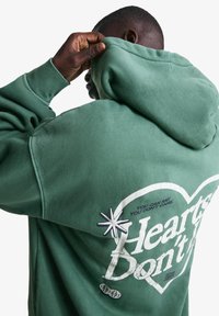 Zelena kapuca s puloverjem iz mehkega blaga. Velik grafični motiv srca z besedilom na hrbtni strani. Oznaka je v belem in vsebuje zvezdico.