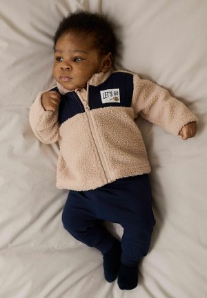 Veste en polaire beige avec des accents bleu marine et un patch "LET'S GO" ; associée à un pantalon bleu marine. Texture douce, fermeture éclair, col rond.
