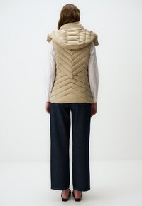 Beige ärmellose Steppweste mit Kapuze und einem Chevron-Steppmuster. Kombiniert mit dunklen, weit geschnittenen Jeanshosen.
