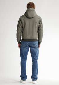 Giacca imbottita verde oliva con cappuccio, orlo e polsini a coste, abbinata a jeans in denim blu e sneakers bianche. Vista posteriore mostrata.