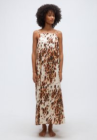 Γυναίκα με σγουρά μαλλιά που φοράει ένα μακρύ, αμάνικο φόρεμα με καφέ και μαύρα στίγματα σε κρεμ φόντο, όρθια και ξυπόλητη.