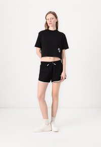 Svart avkortet t-skjorte med logo på venstre side, kombinert med svarte shorts med hvit snor og kantdetaljer. Brukt med hvite joggesko.