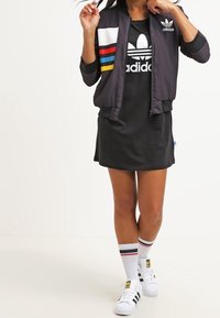 Abito nero a maniche corte, abbinato a un bomber cropped con zip e strisce colorate. Indossato con calzini bianchi e sneakers.