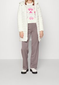 Weißer Daunenmantel mit Kapuze, über einem cremefarbenen Pullover mit rosa und grauen Herzmustern, kombiniert mit grauen weiten Hosen und weißen Stiefeln.