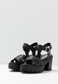 Czarne sandały na platformie z masywnym obcasem, z krzyżującymi się paskami, regulowanym zapięciem i teksturowaną podeszwą zapewniającą przyczepność.