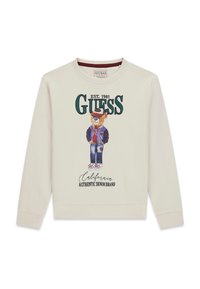 Krémová mikina s potiskem kresleného medvěda v varsity bundě, s kšiltovkou a džínami; text zní "GUESS EST. 1981 California Authentic Denim Brand."