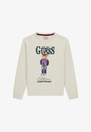 Krémová mikina s potiskem kresleného medvěda v varsity bundě, s kšiltovkou a džínami; text zní "GUESS EST. 1981 California Authentic Denim Brand."