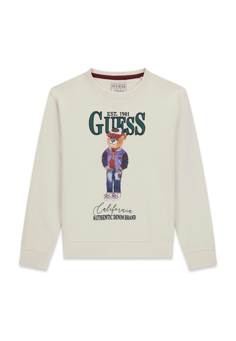 Krémová mikina s potiskem kresleného medvěda v varsity bundě, s kšiltovkou a džínami; text zní "GUESS EST. 1981 California Authentic Denim Brand."