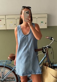 Vaaleansininen farkku hihaton mekko, jossa on pyöreä pääntie ja etusaumadetail, jota on käytetty ruskean vyön kanssa polkupyörän edessä.