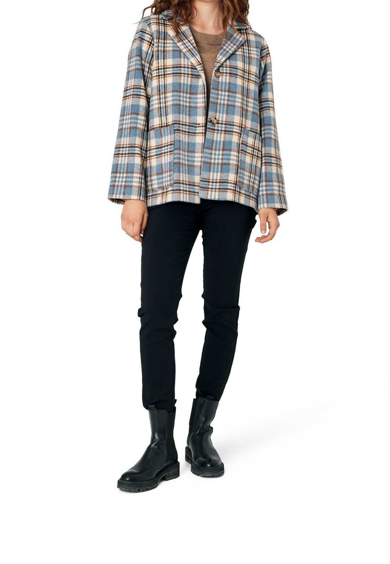 Femme portant une veste à carreaux bleue et beige sur un top marron, un pantalon slim noir et des bottines en cuir noir, debout sur un fond blanc.