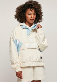 Lichte crème windjack met blauwe accenten, elastische manchetten en een oversized ontwerp. Kenmerkt zich door een prominent "STARTER" logo op de voorkant.