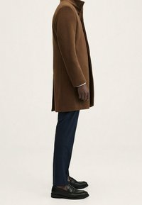 Uomo in soprabito marrone, pantaloni blu navy e mocassini neri in piedi di lato su uno sfondo neutro.