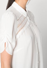 White blouse featuring a collared neck, sheer lace insert on the chest with decorative patterns, and puffed short sleeves with gathered detail.