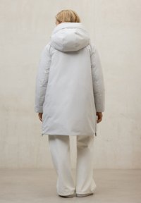 Abrigo largo acolchado con capucha en gris claro, con cierre frontal con cremallera, tejido texturizado y cremalleras laterales. Se lleva sobre pantalones blancos.