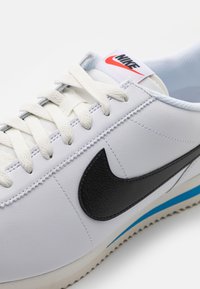 Fehér bőrszövetű sneaker fekete Swoosh logóval, texturált díszítésekkel, krémszínű fűzőkkel és kék csíkkal a talpán. Piros Nike márkajegyzés a nyelvén.