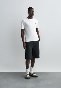 White t-shirt with small orange graphic, paired with black shorts. Model wears white socks and beige shoes with dark soles. Neutral background.