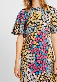 Frau trägt ein knielanges schwarzes Kleid mit mehrfarbigen Blumenmustern, puffärmeligen kurzen Ärmeln und einem Schlitz vorne.