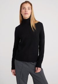 Schwarzer Rollkragenpullover aus weichem Strickmaterial mit gerippten Bündchen und Saum, kombiniert mit grauen maßgeschneiderten Hosen. Schlichtes, modernes Design.