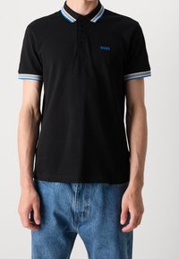 Homme portant un polo noir avec un bord bleu et blanc au col et aux manches, logo "BOSS" bleu sur la poitrine, associé à un jean bleu.