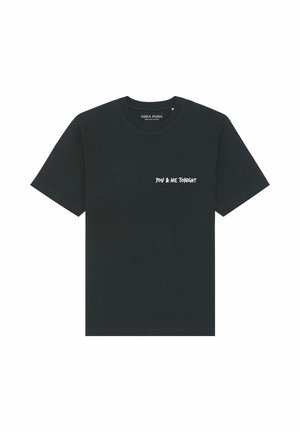 Jeune femme aux cheveux bruns et lisses portant un T-shirt noir oversize avec le texte blanc « YOU & ME TONIGHT » et un jean bleu clair.