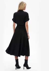 Schwarzes, knielanges Kleid mit kurzen Ärmeln, plissiertem Rock und taillierter Taille. Kombiniert mit schwarzen, knöchelhohen Stiefeln mit Absatz, aus glattem Stoff, ohne sichtbare Muster.