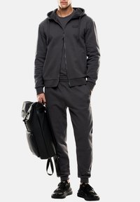 Tuta grigia scura con felpa con zip e pantaloni joggers, realizzata in tessuto morbido, abbinata a uno zaino nero e sneakers nere.