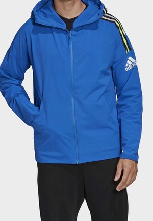 Homme portant une veste à capuche Adidas bleue avec fermeture éclair frontale et pantalon noir, mains détendues, sur fond uni.