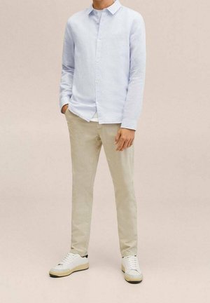 Mężczyzna w jasnoniebieskiej koszuli na guziki, beżowych spodniach i białych sportowych butach, stoi z jedną ręką w kieszeni na jednolitym tle.