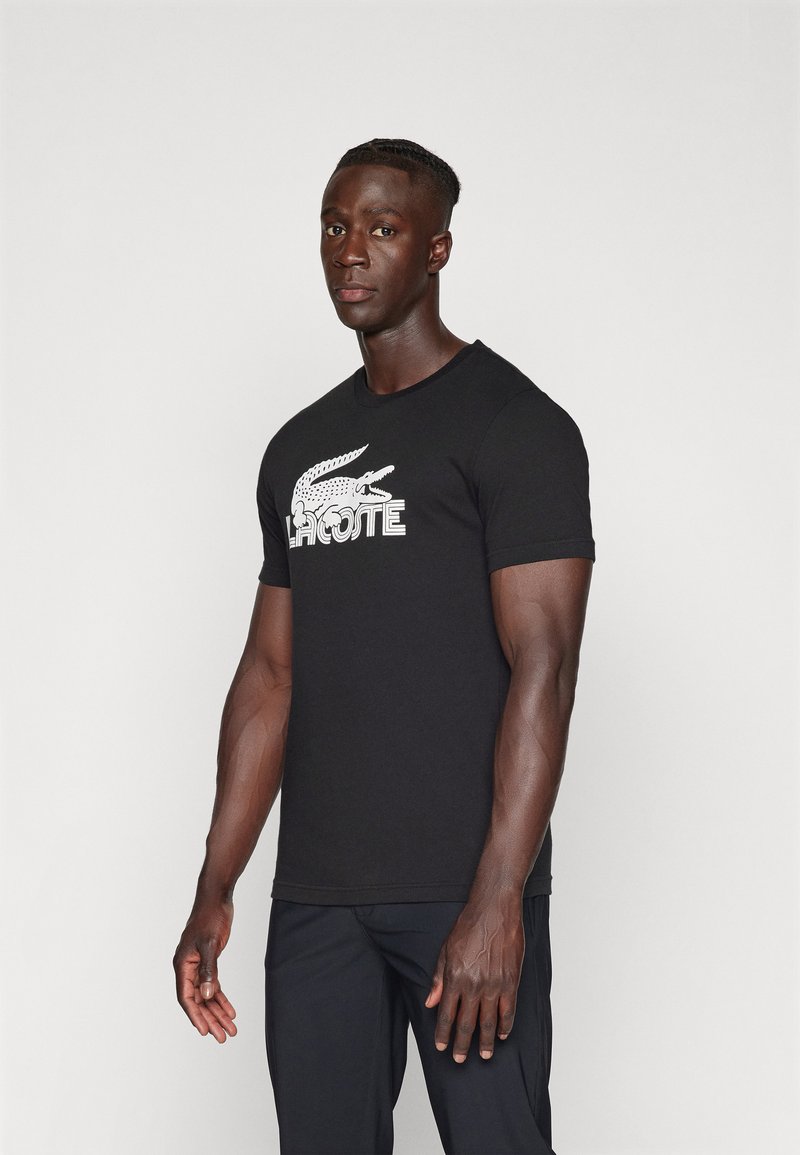 Camiseta de algodón negra con un logo de cocodrilo blanco y el texto "LACOSTE", de forma ajustada y con mangas cortas. Textura suave.