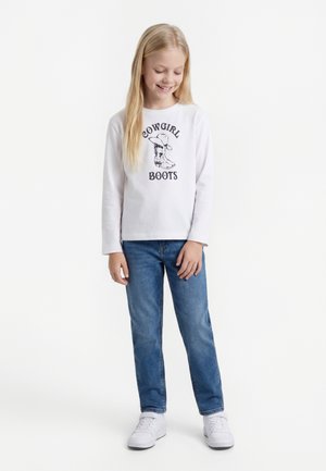 Dziewczyna z długimi blond włosami, ubrana w białą koszulkę "Cowgirl Boots", niebieskie dżinsy i białe trampki, stoi i uśmiecha się z zamkniętymi oczami.