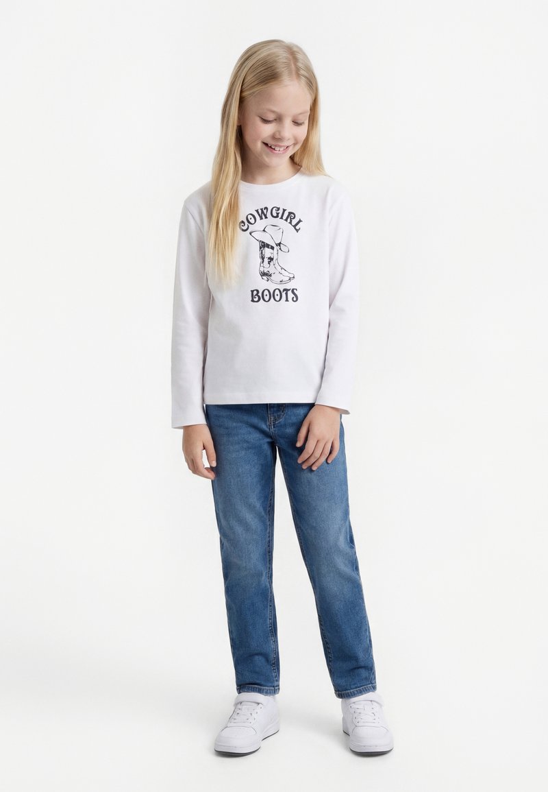 Tüdruk, kellel on pikad blondid juuksed, seljas valge „Cowgirl Boots“ särk, sinised teksad ja valged tossud, seisab ja naeratab silmad kinni.